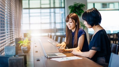 Two women sat at a desk looking at a tablet and laptop in a modern office.