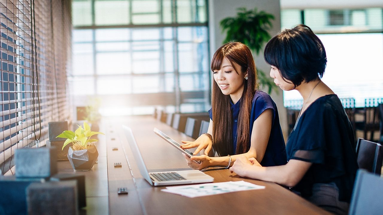 Two women sat at a desk looking at a tablet and laptop in a modern office.