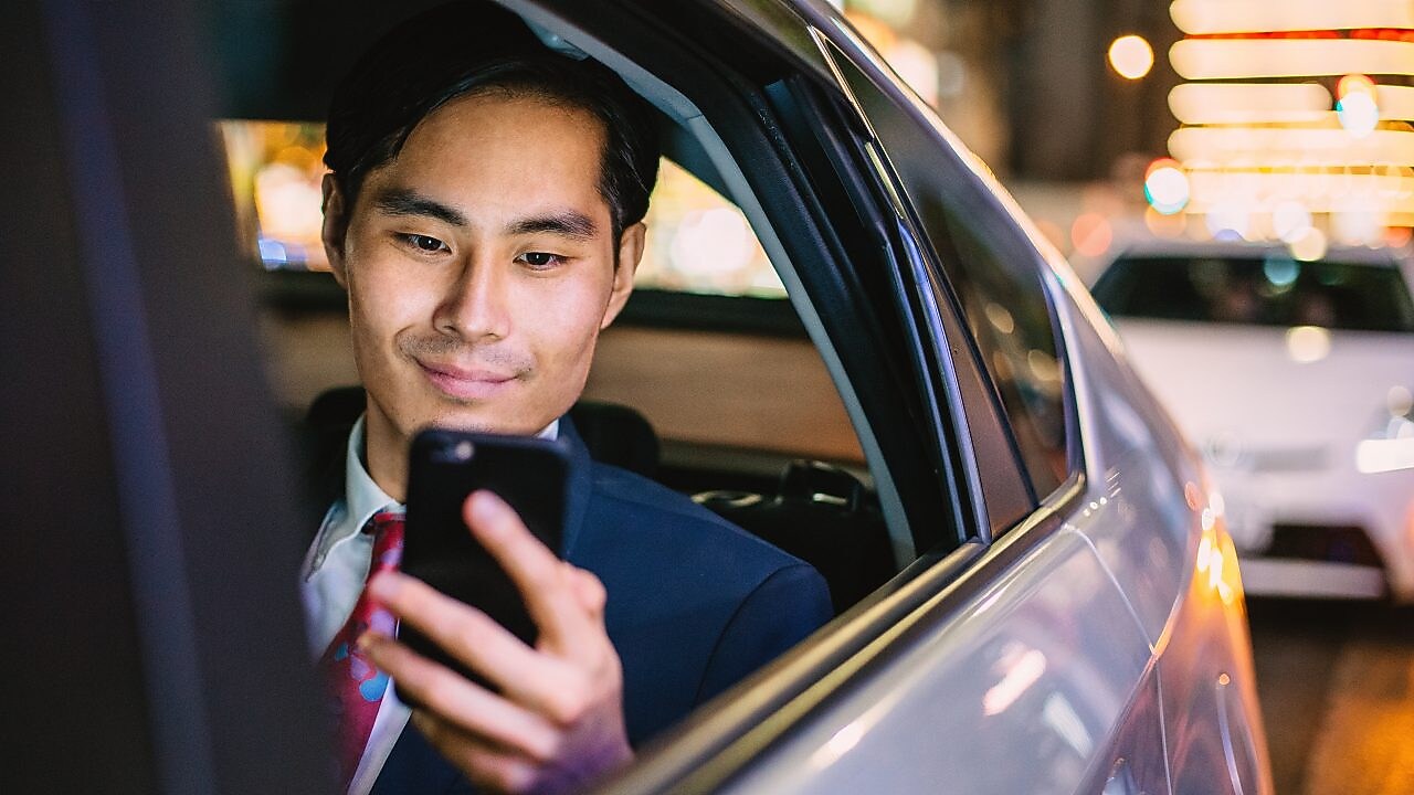 image of a man through the open backseat window of a car while he looks at his mobile device.
