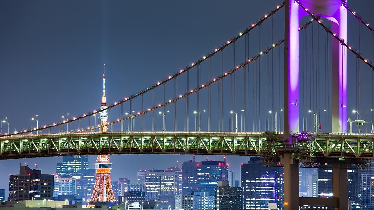 a night time image of the Rainbow bridge in Japan with Tokyo Tower in the background. The bridge is lit up with green and purple lighting.