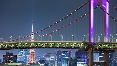 a night time image of the Rainbow bridge in Japan with Tokyo Tower in the background. The bridge is lit up with green and purple lighting.
